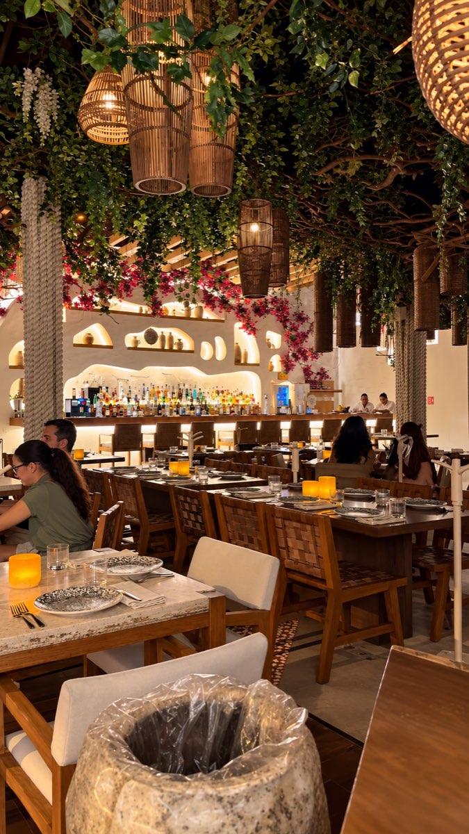 A restaurant interior with a leafy canopy, warm woven lights, and dining tables